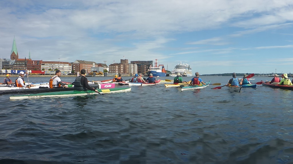 Viele Kajaks auf der Kieler Förde. Im Hintergrund sieht man den Kieler Rathausturm und ein Kreuzfahrtschiff