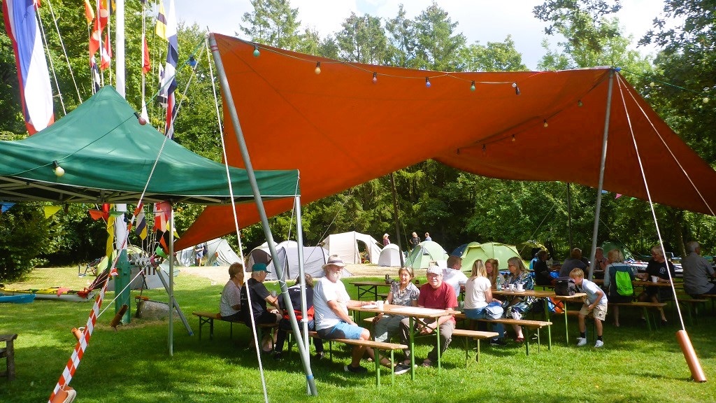 Teilnehmende sitzen an Bierzeltgarnituren unter dem Sonnensegel auf der Wiese vom TSV Klausdorf